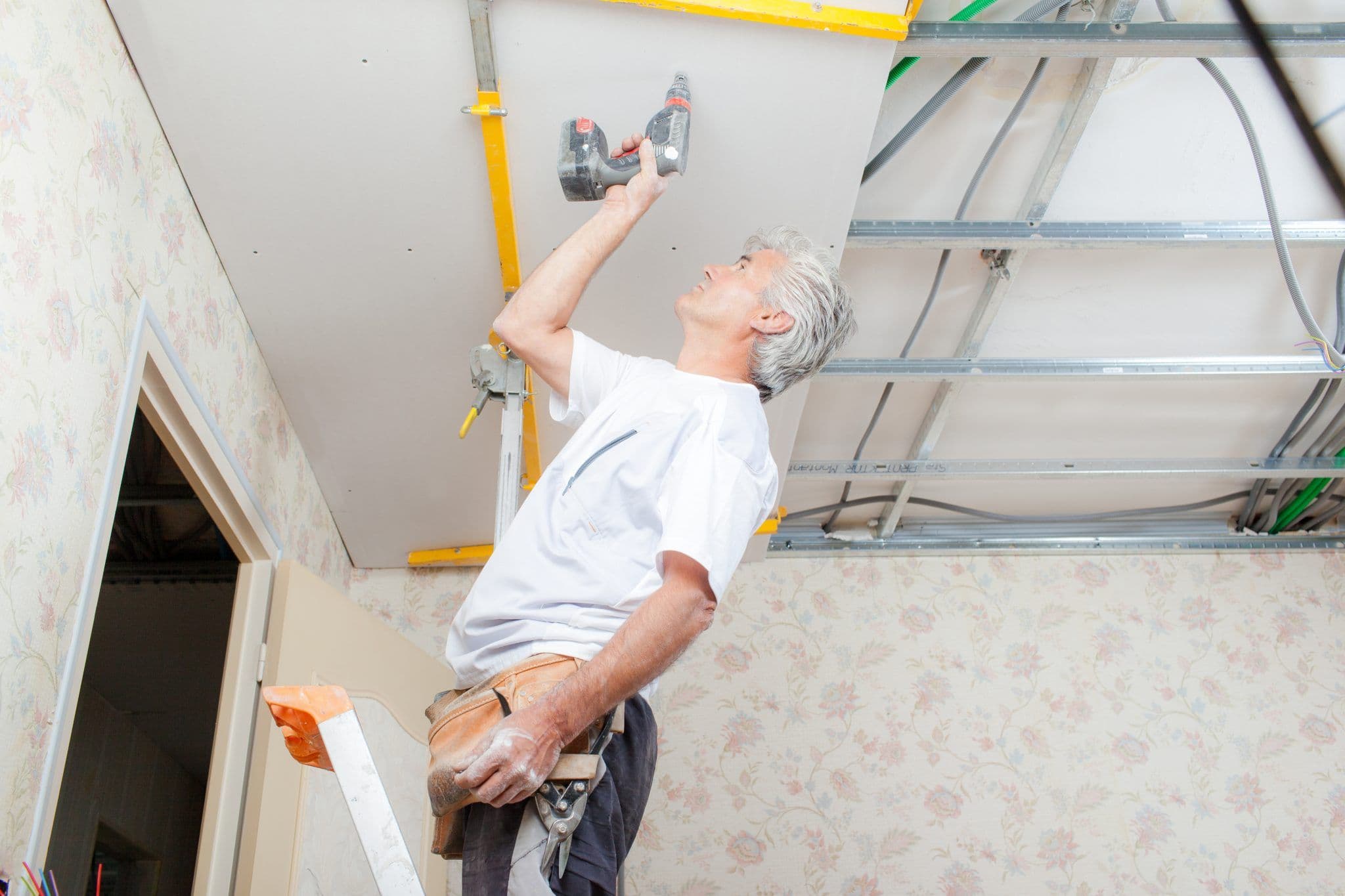 Image of an older man on a ladder installing drywall on a ceiling with a power drill, inside a room under renovation.
