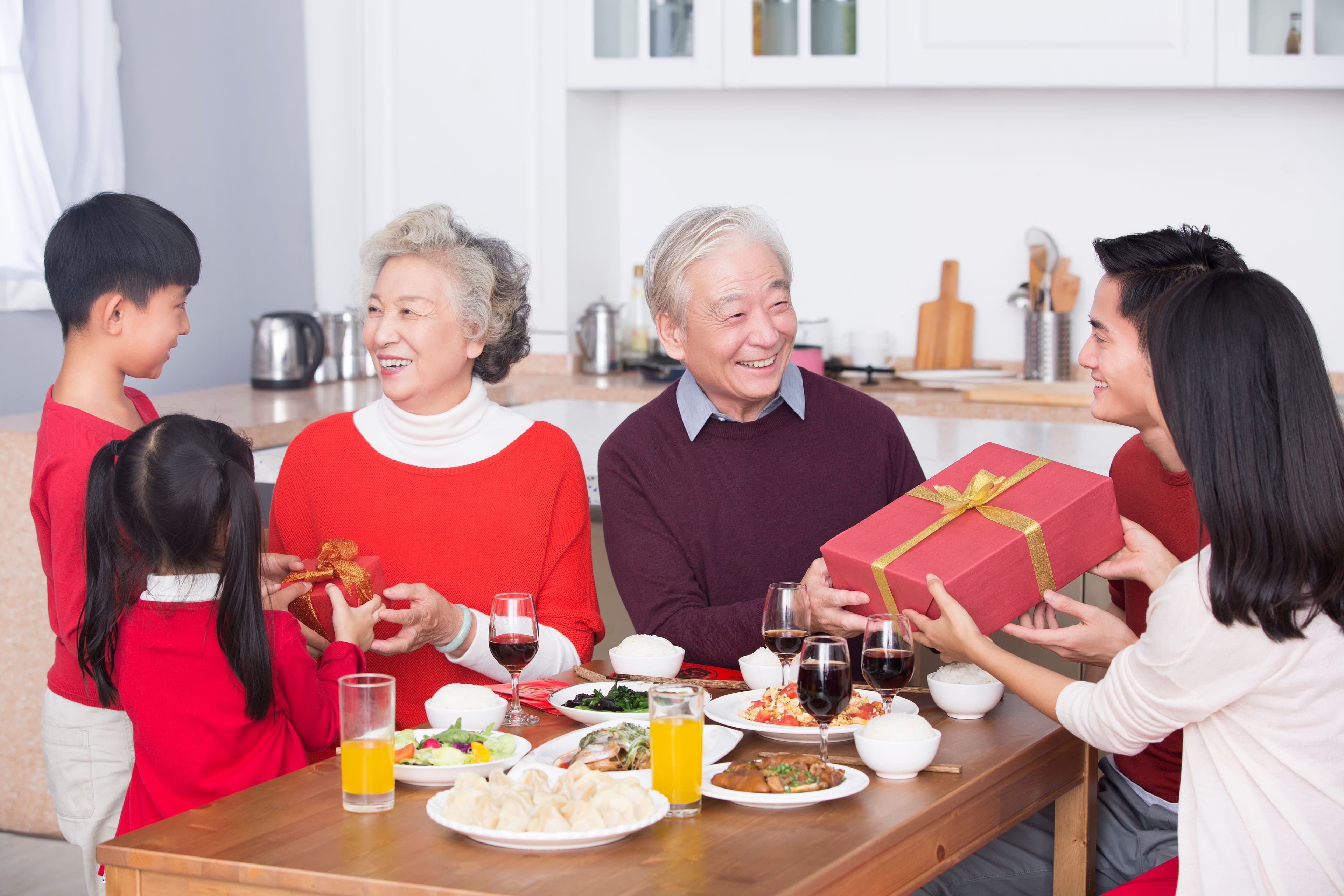 Image of a multi-generational Asian family gathered around a dinner table, joyfully exchanging gifts and sharing a festive meal together.