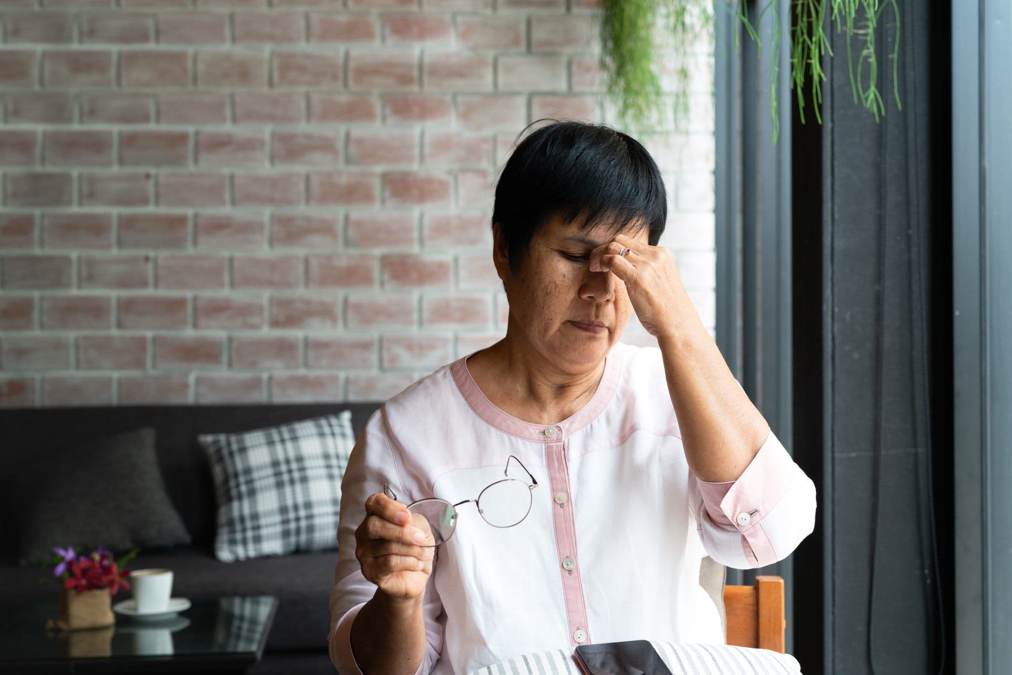 Image of an older woman sitting indoors, holding her glasses and rubbing her forehead, appearing tired or stressed.