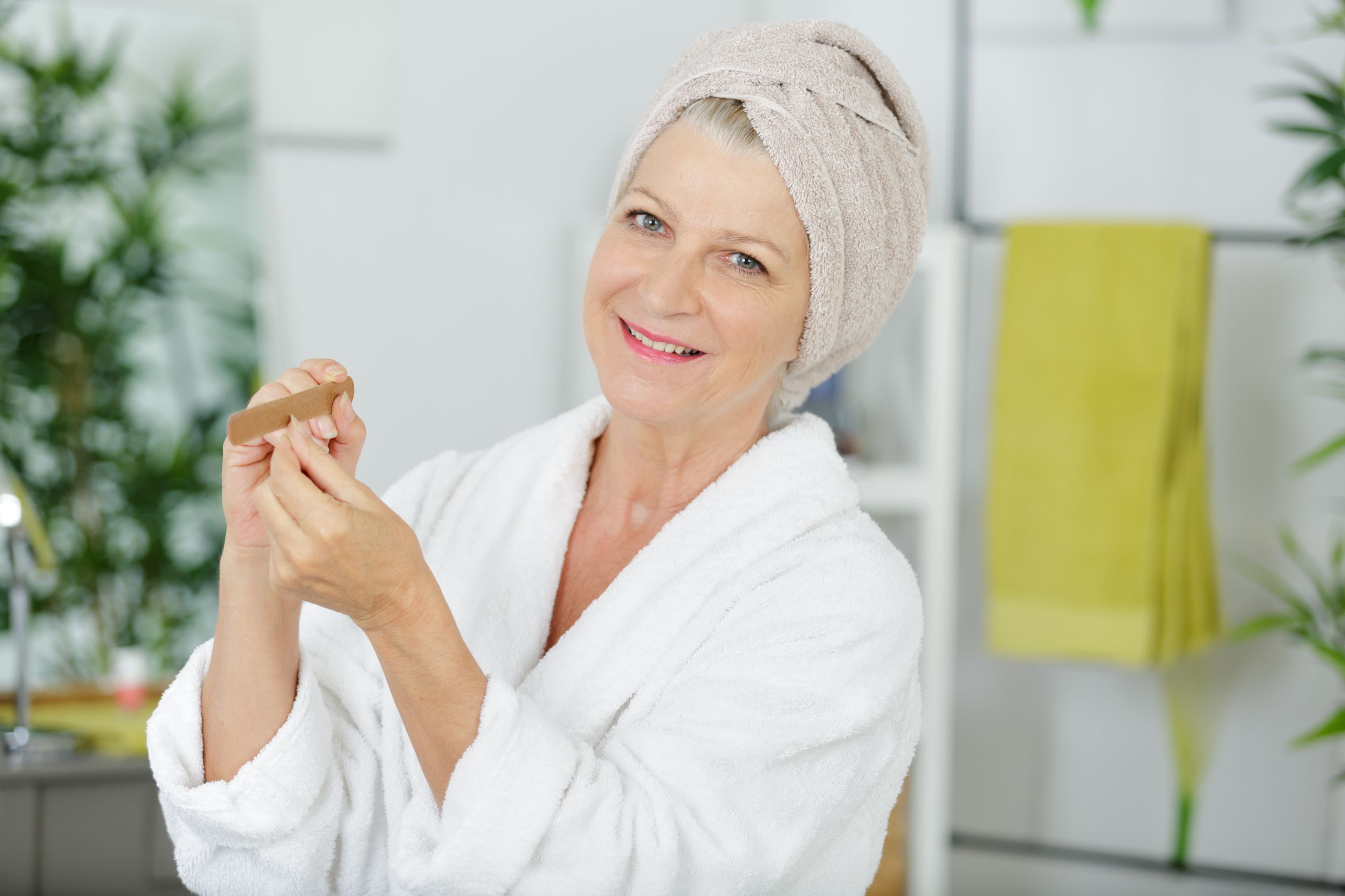 Image of an Smiling older woman in a white bathrobe and towel filing her nails in a bright, modern bathroom.